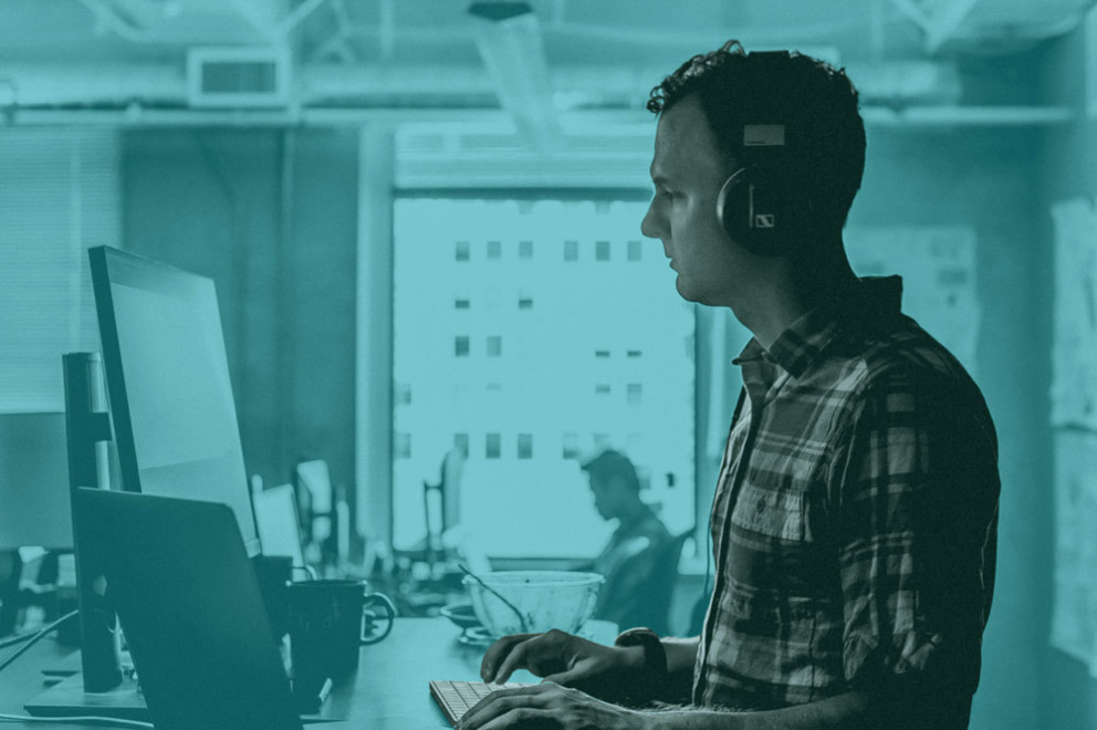 Cyan duotone man standing at computer wearing headphones.